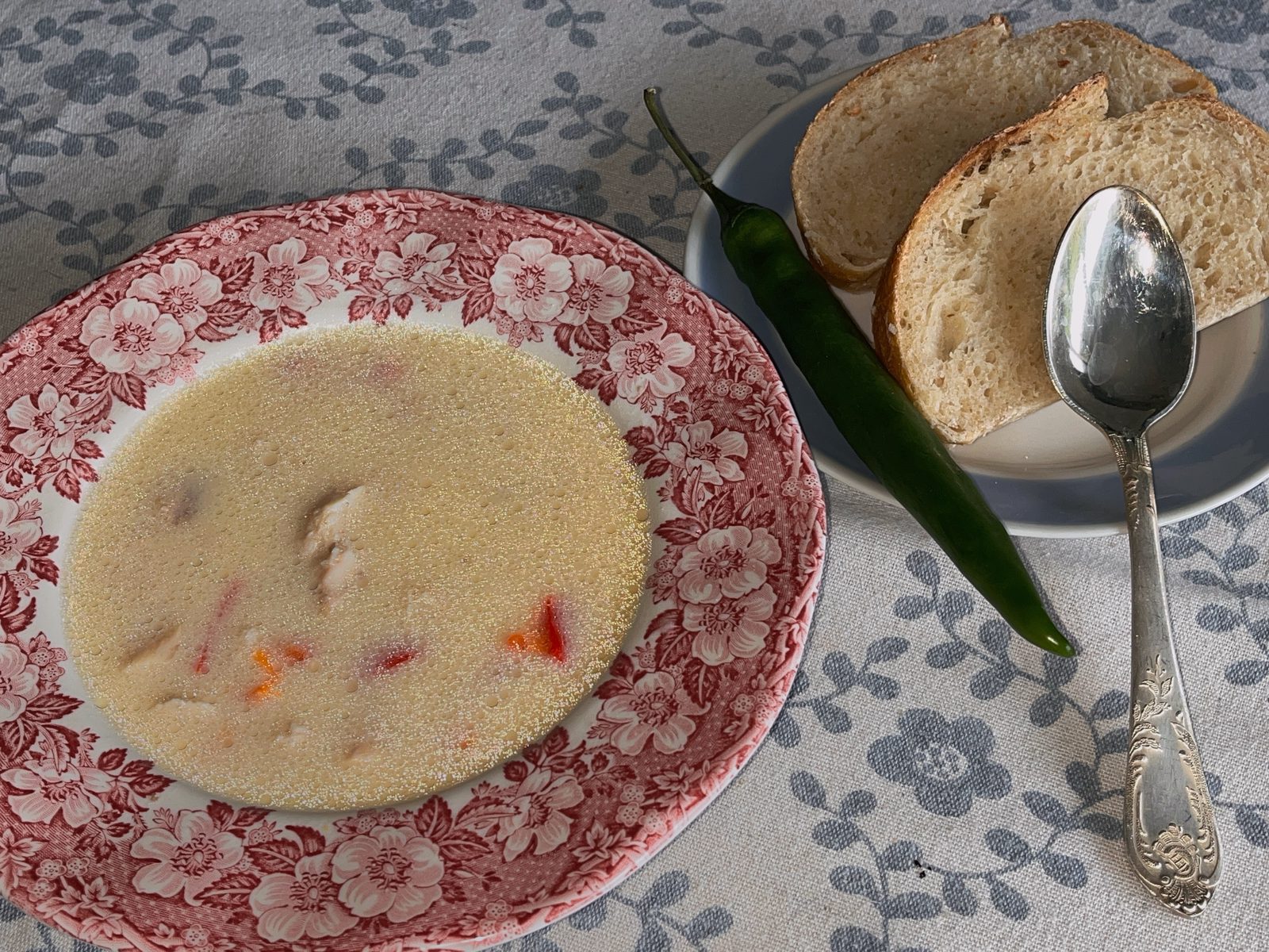 A bowl of soup, served with bread and a green chili pepper.