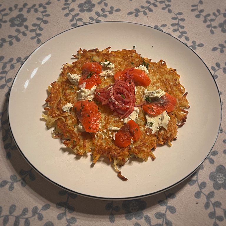A large round hash brown on a plate, served with cured fish, cream cheese and pickled red onion.