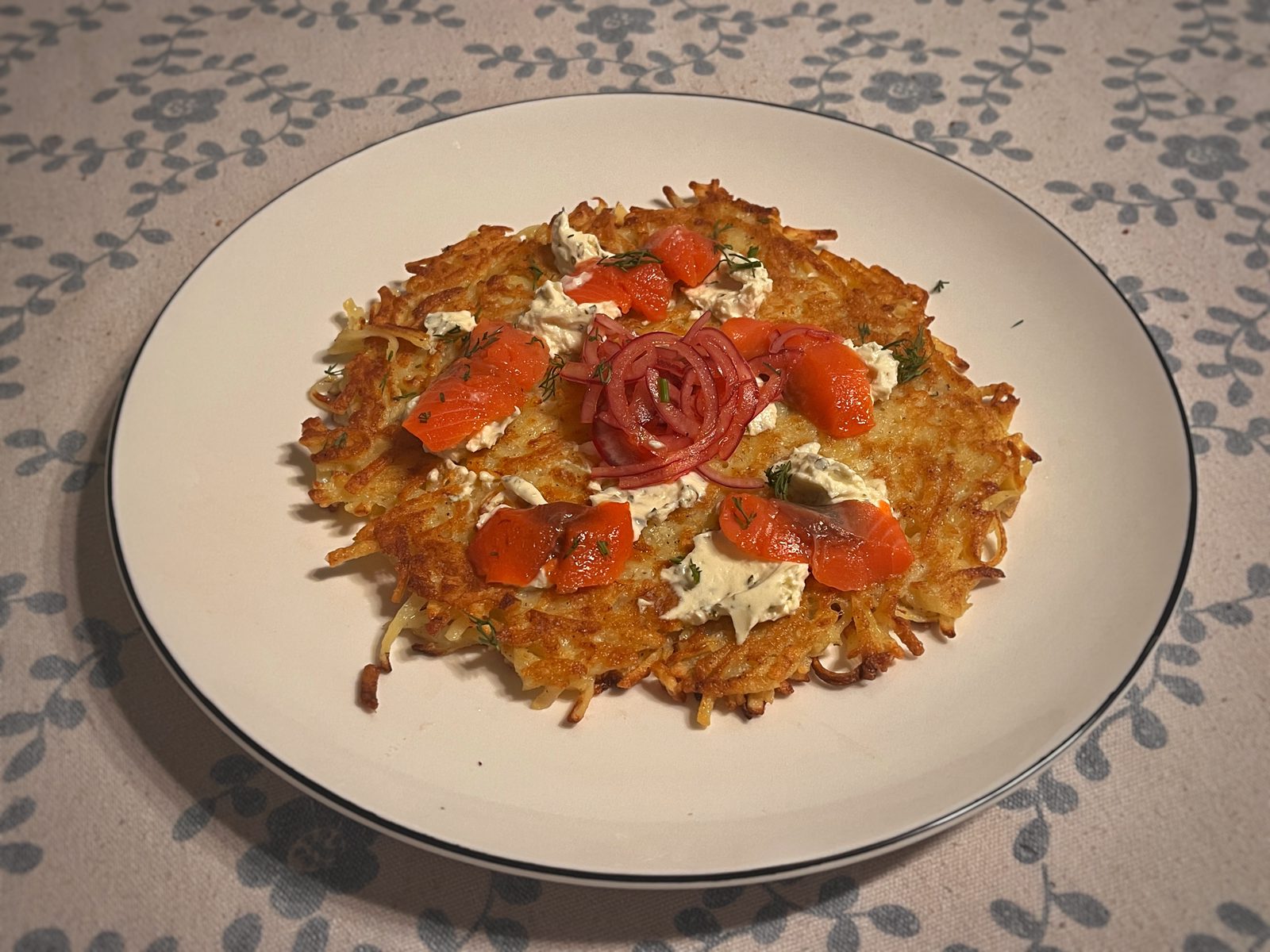A large round hash brown on a plate, served with cured fish, cream cheese and pickled red onion.